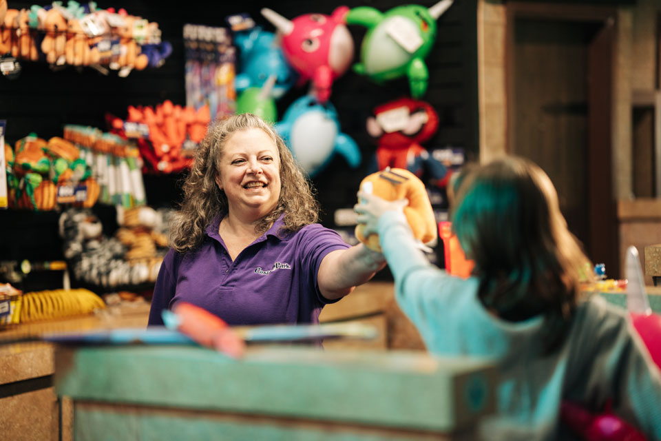 A woman in a purple shirt smiles as she hands a plush toy to a child across a counter in a toy store with colorful stuffed animals in the background.