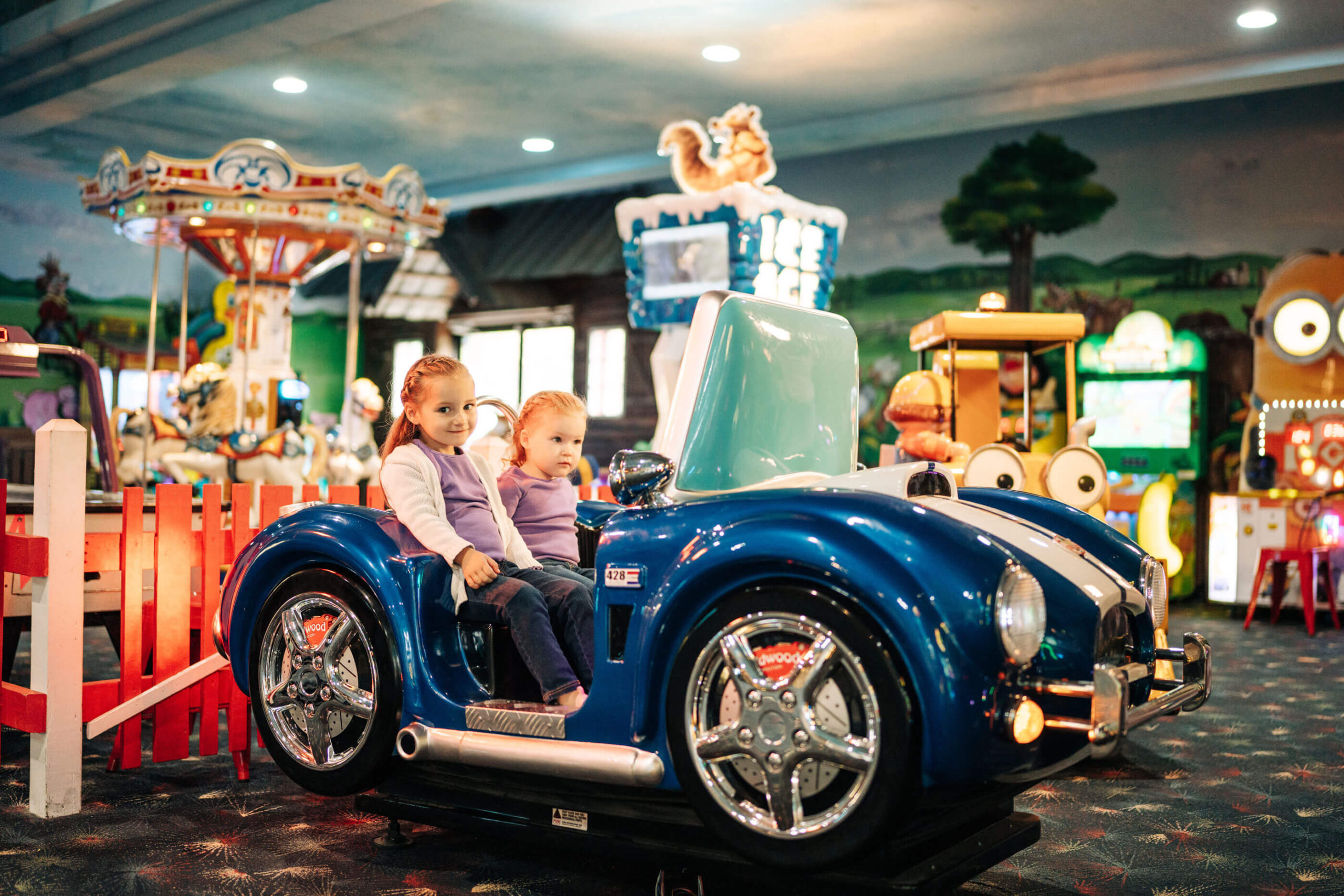Two young children sit in a blue toy car in an indoor amusement area. The girl on the left wears a purple sweater, and the boy on the right wears a gray sweater. Colorful rides and games are visible in the background.
