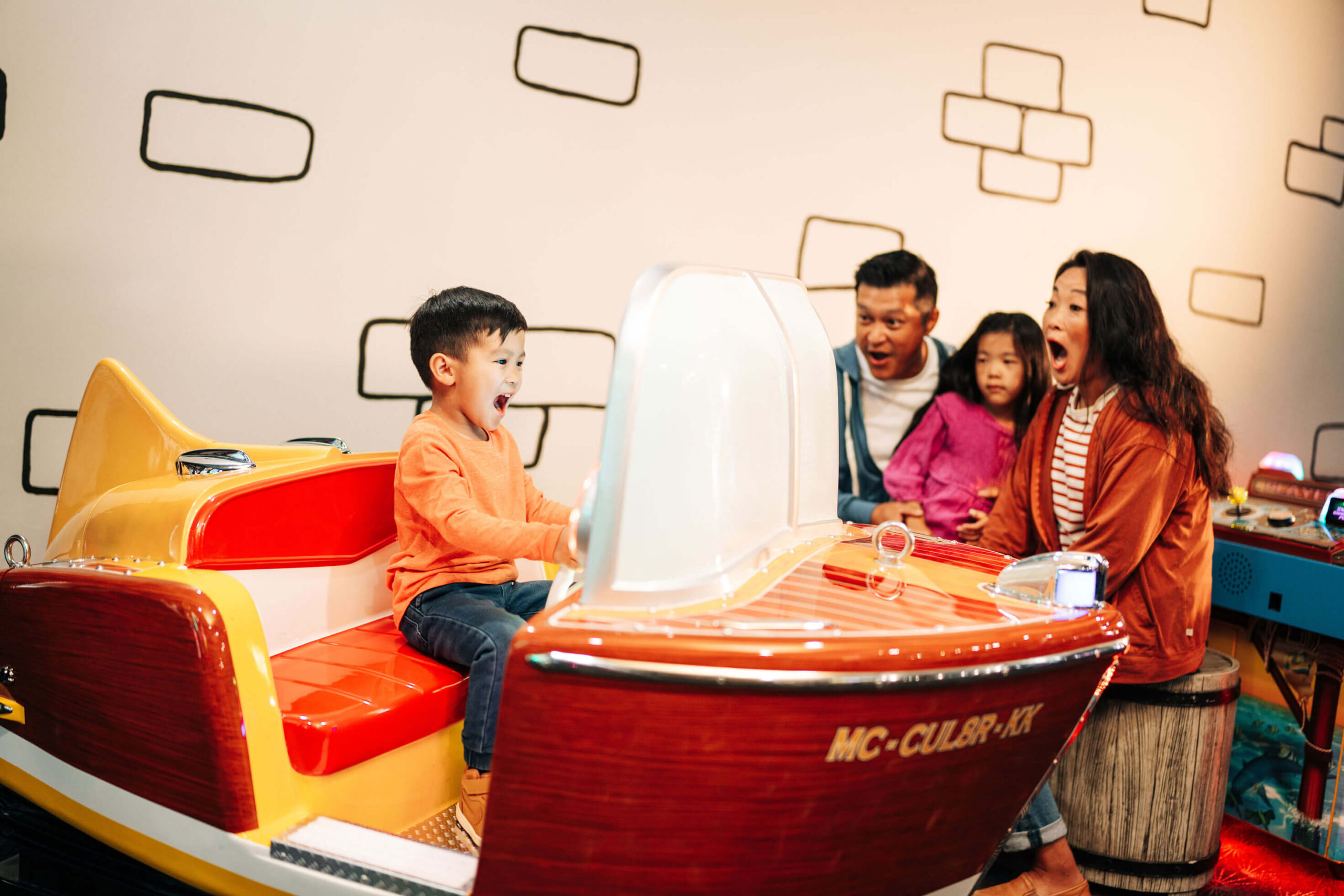 A child excitedly rides a boat-shaped amusement ride, while two adults and another child watch with joy. The room has a white wall with black line designs. The ride is brightly colored with "MC-6U8P-XX" on it.