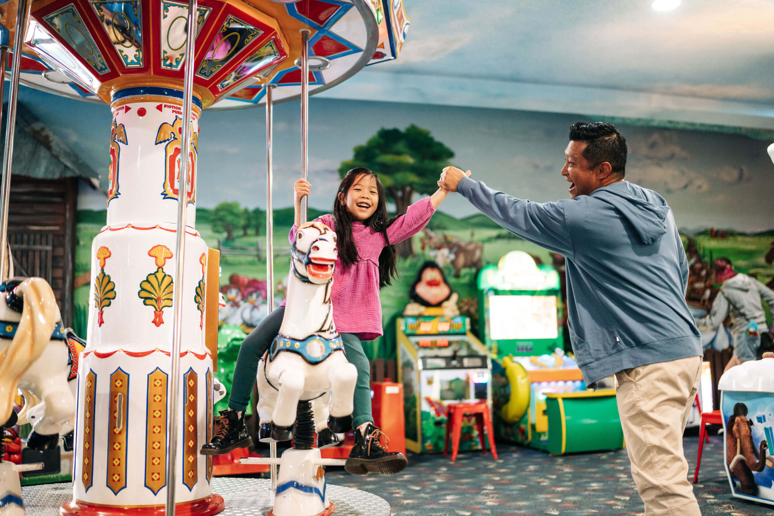 A young girl in a pink sweater enjoys a merry-go-round ride, smiling and holding hands with a man in a blue jacket. They are in a colorful indoor amusement area with various rides and games in the background.