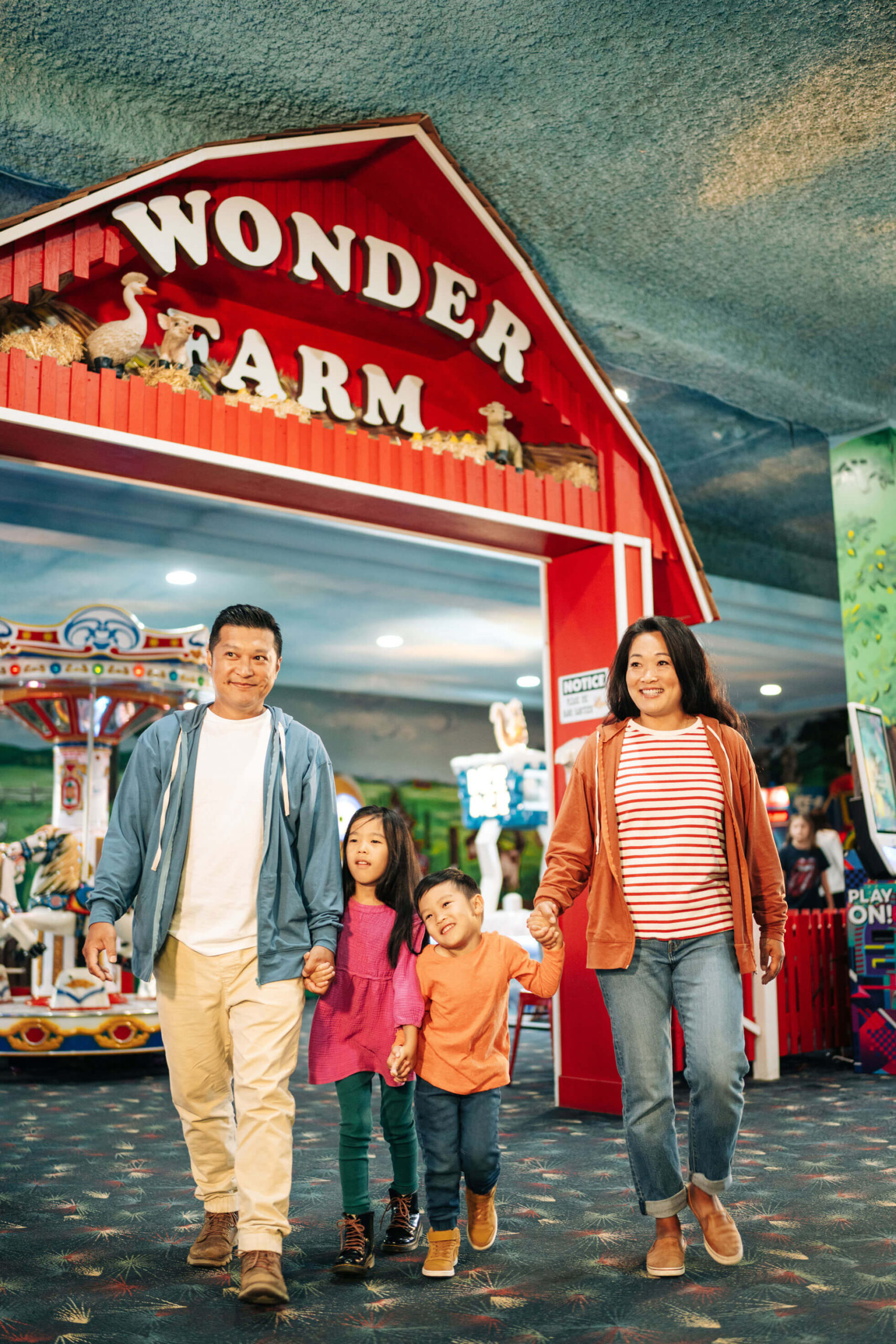 A smiling family of four walks hand in hand at an amusement park. They are in front of a red building labeled "Wonder Farm" with a carousel in the background. The setting looks vibrant and cheerful.