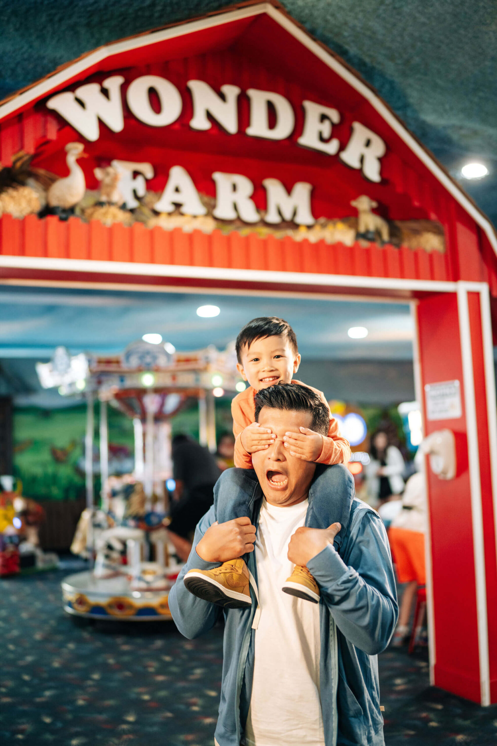 A man playfully carries a child on his shoulders, with the child covering the man's eyes. They are smiling and standing under a red "Wonder Farm" sign in an indoor amusement area with colorful lights and rides in the background.