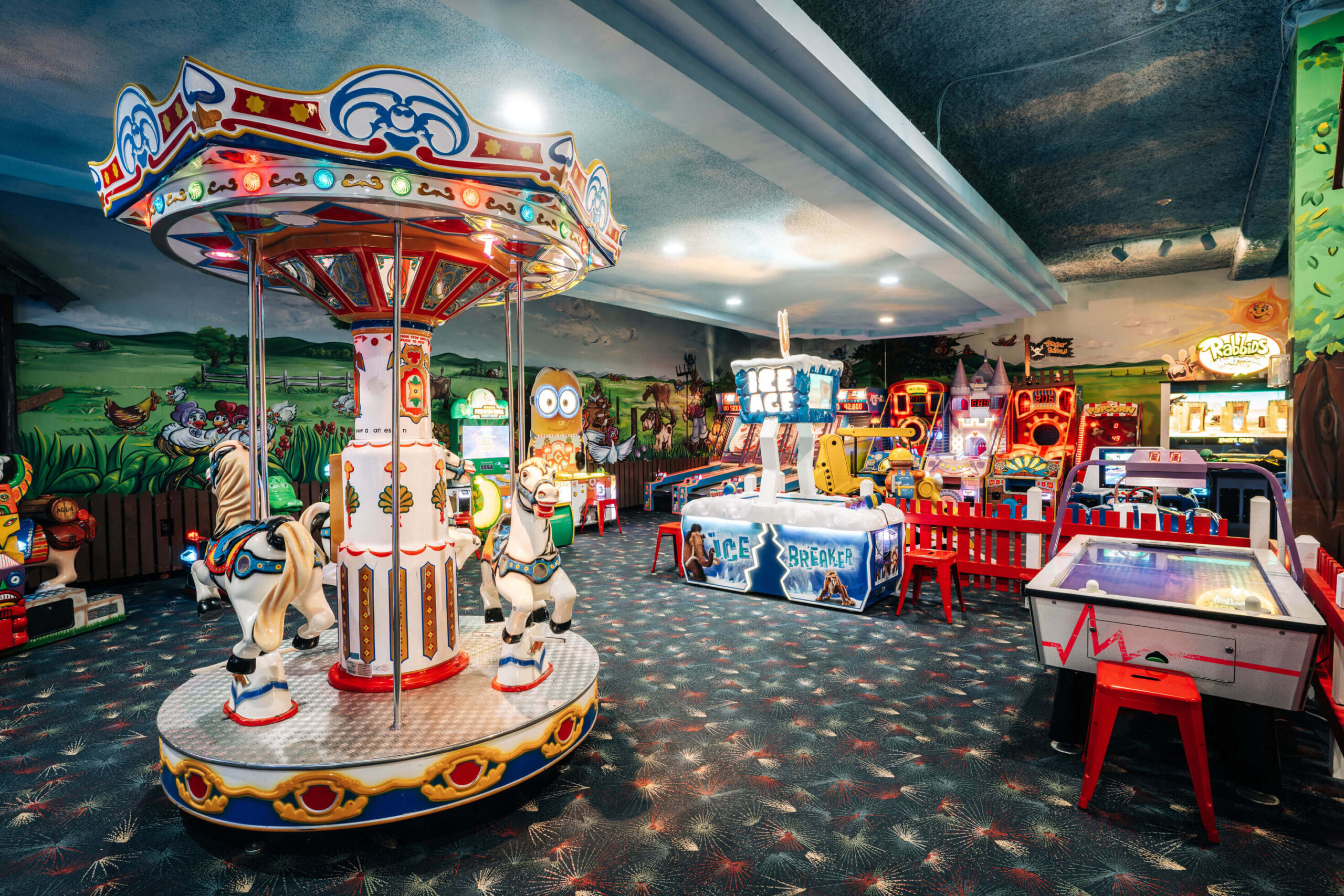 A colorful arcade with a small carousel, bright machines, and a variety of games. The carpet features a star pattern, and the walls are painted with outdoor scenes. The atmosphere is vibrant and playful.