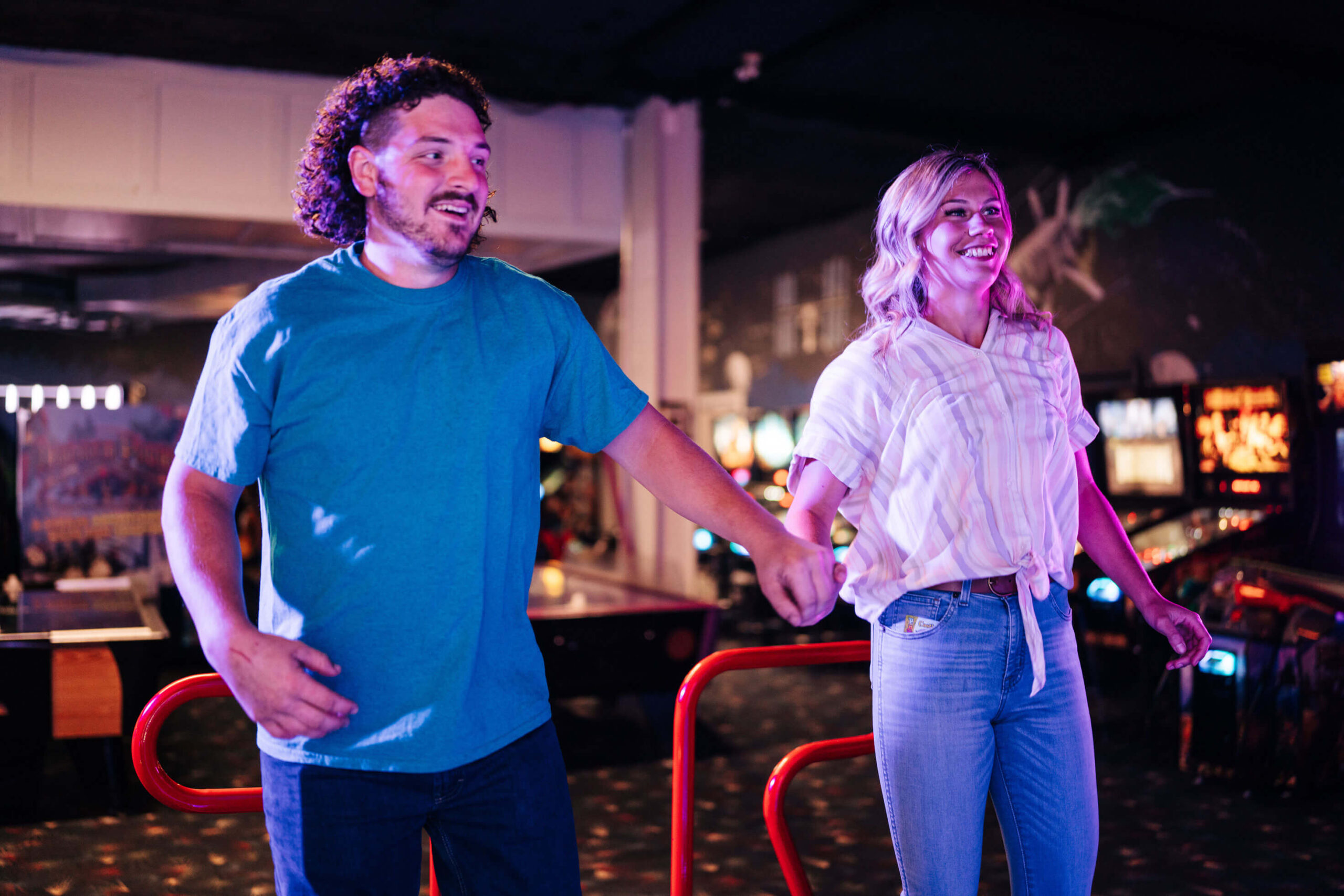 A man and a woman are smiling and holding hands while playing at an arcade. They appear to be stepping on a dance game platform, surrounded by colorful arcade machines and lights.