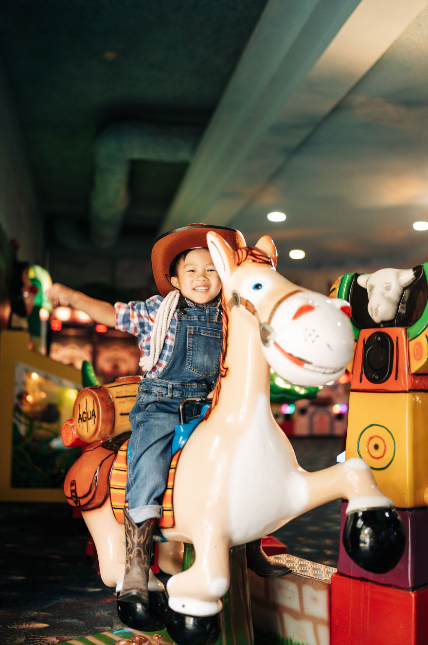A cheerful child in overalls and a cowboy hat rides a mechanical horse in an amusement arcade. The background is colorful with various lights and arcade games, creating a lively atmosphere.