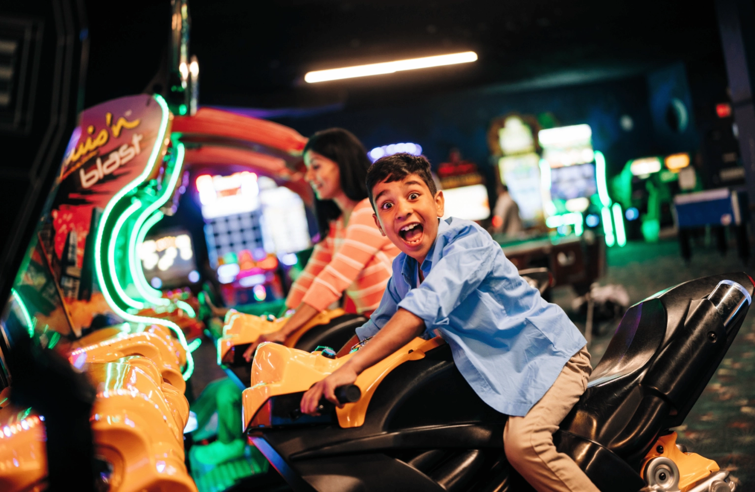 A young boy excitedly rides an arcade motorcycle game, smiling widely, while a girl in the background plays a similar game in a brightly lit arcade with colorful neon lights.