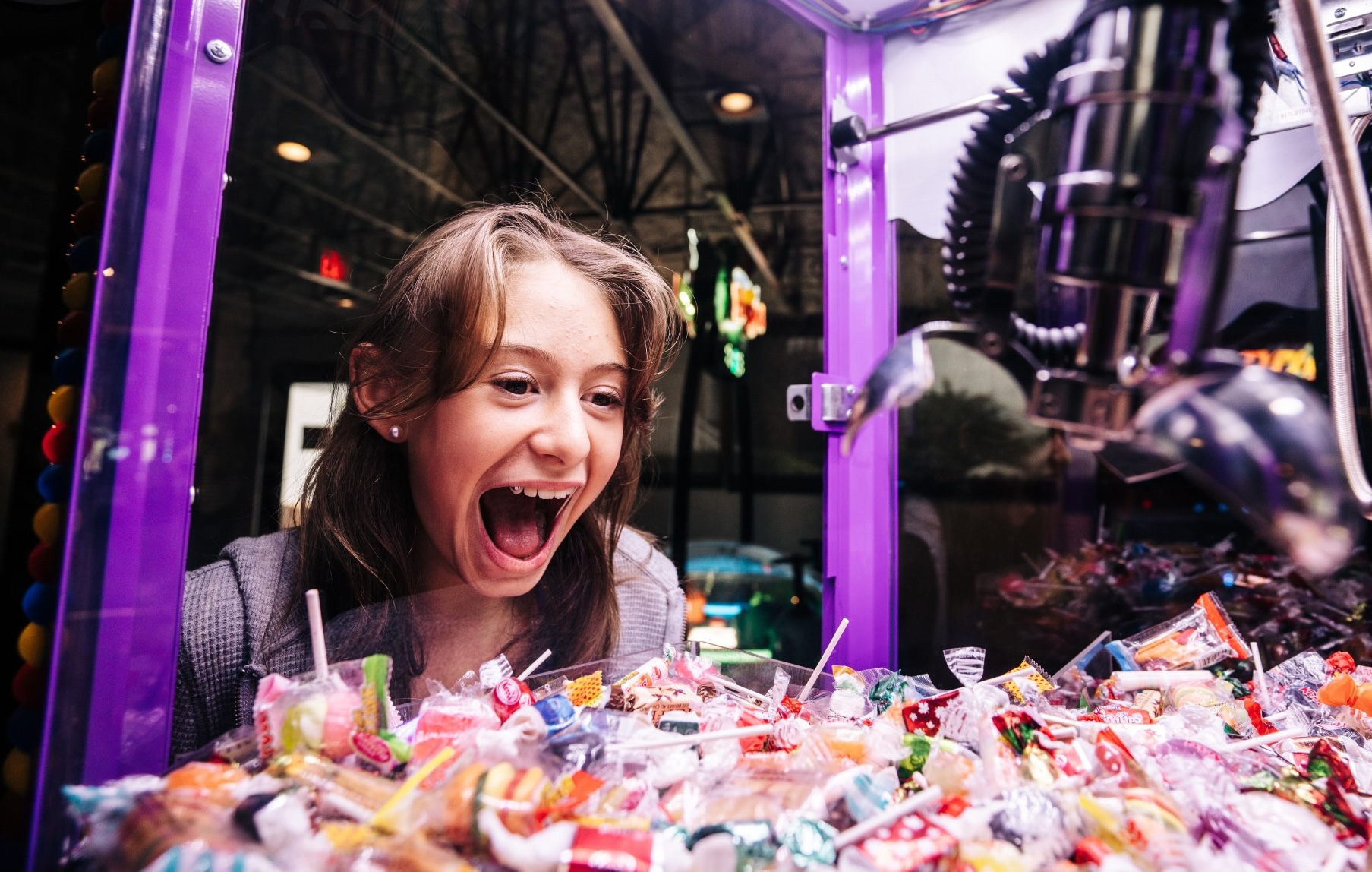 A young girl excitedly looks into a claw machine filled with colorful candy and treats, her mouth open in delight as she eagerly watches the claw descend.