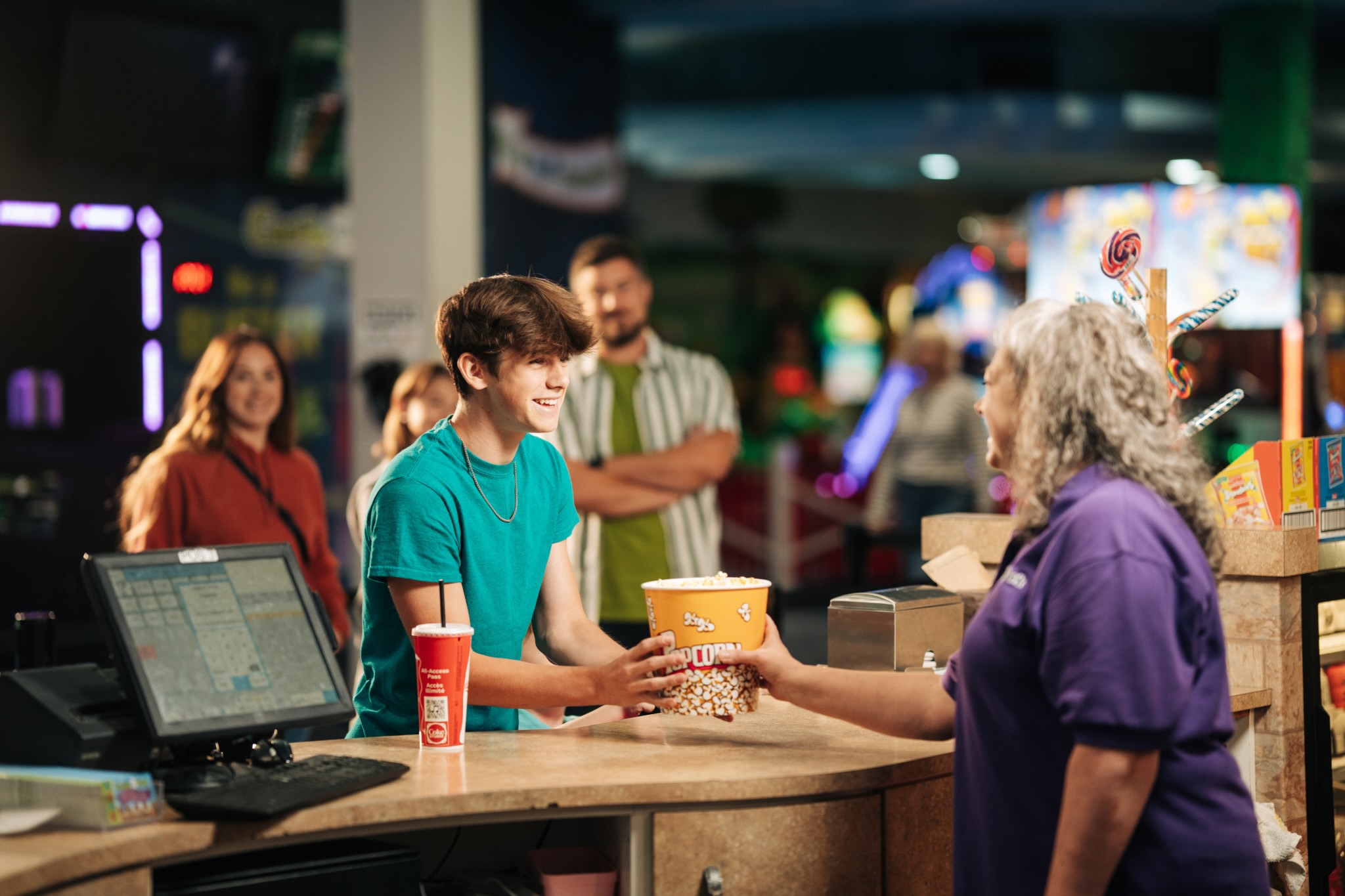 A young man at a concession stand in an arcade smiles as he receives popcorn from a staff member in a purple shirt; other people wait in line and arcade games are visible in the background.