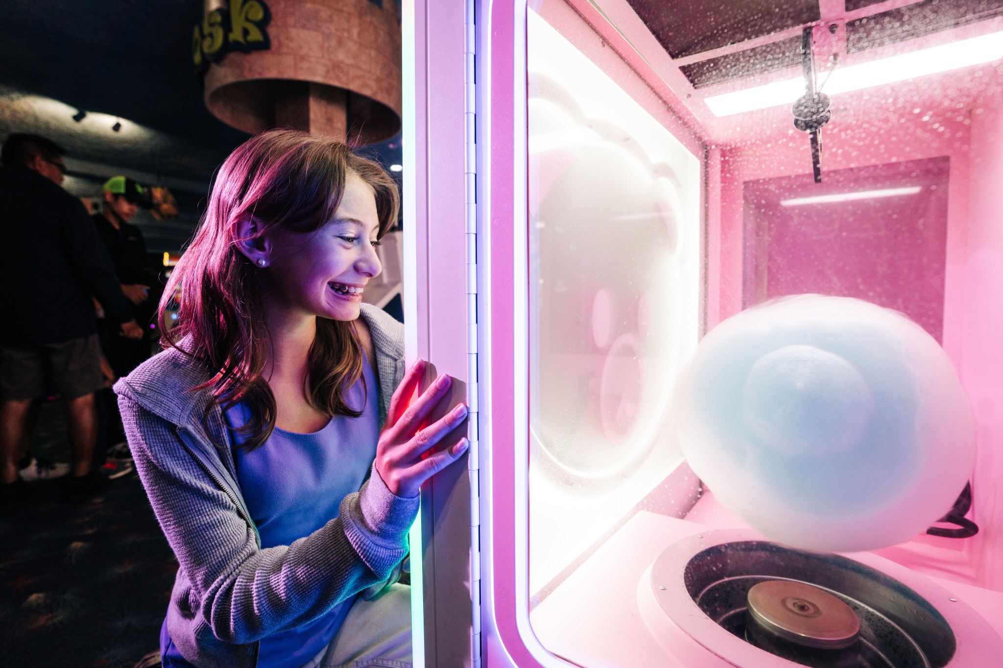 A young woman smiles excitedly while watching a cotton candy machine spin blue cotton candy inside a brightly lit arcade. Neon pink and purple lights glow around her.