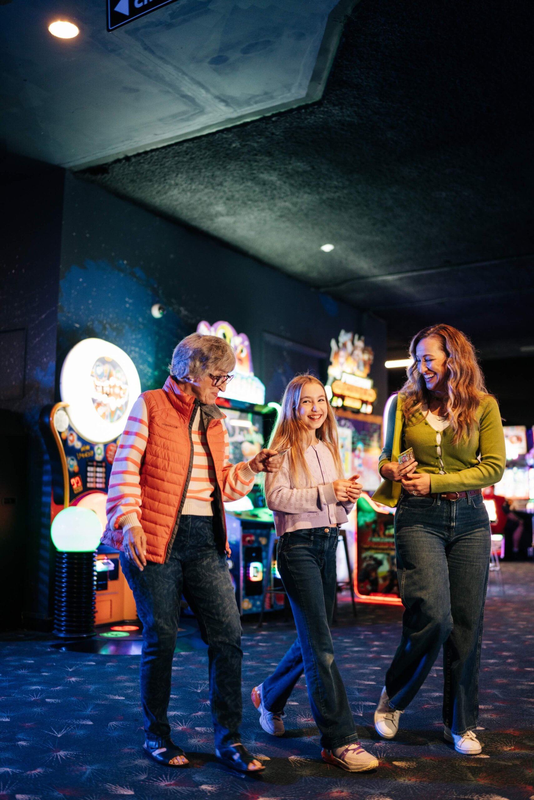 Three generations of women—an older woman, a young girl, and an adult woman—smile and walk together inside an arcade filled with colorful lights and games. They look happy and are holding hands.